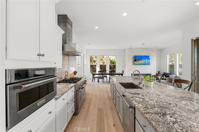 kitchen with white cabinets, appliances with stainless steel finishes, light stone countertops, wall chimney range hood, and a sink