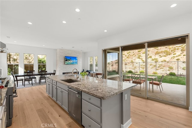 kitchen featuring gray cabinets, stainless steel dishwasher, a kitchen island with sink, a sink, and light stone countertops