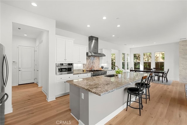 kitchen featuring a kitchen island with sink, stainless steel appliances, a sink, white cabinets, and wall chimney range hood