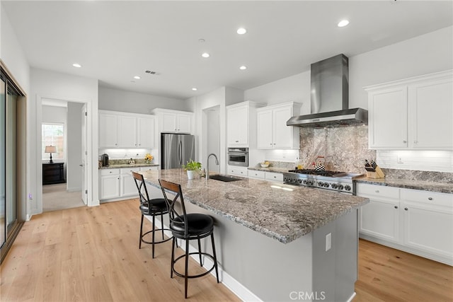 kitchen featuring appliances with stainless steel finishes, white cabinets, a kitchen island with sink, a sink, and wall chimney range hood