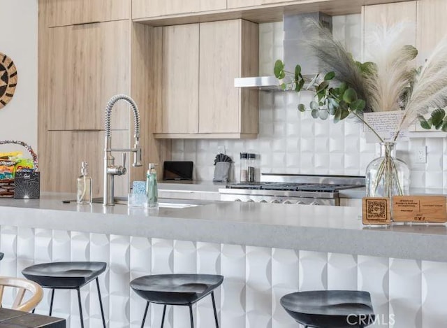 kitchen with tasteful backsplash, a breakfast bar area, light brown cabinets, light stone counters, and wall chimney range hood