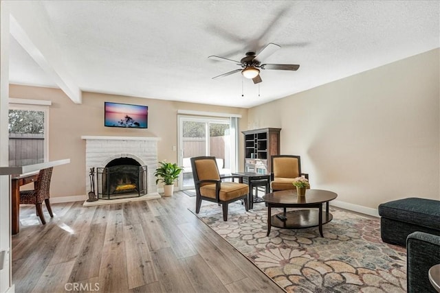 living room featuring beamed ceiling, ceiling fan, light hardwood / wood-style floors, a brick fireplace, and a textured ceiling