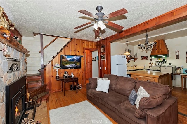 living room with ceiling fan with notable chandelier, wooden walls, a textured ceiling, a stone fireplace, and light wood-type flooring