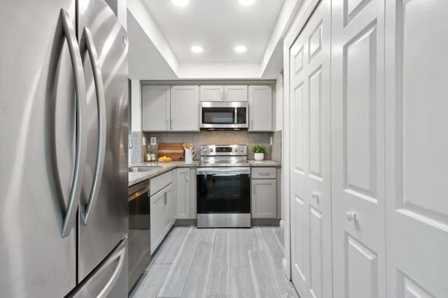 kitchen featuring light hardwood / wood-style flooring, a tray ceiling, gray cabinets, stainless steel appliances, and backsplash