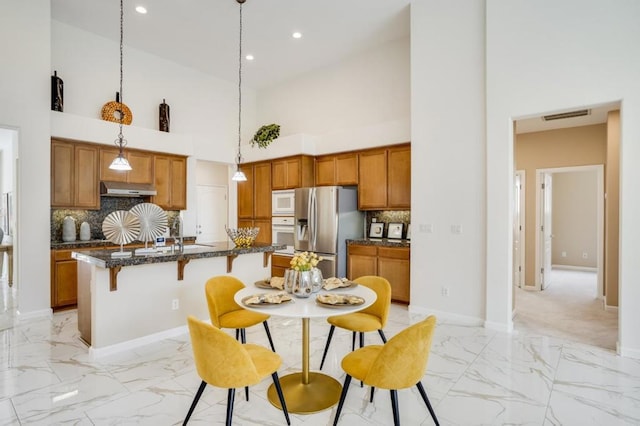 kitchen featuring decorative light fixtures, tasteful backsplash, a high ceiling, a center island with sink, and white appliances