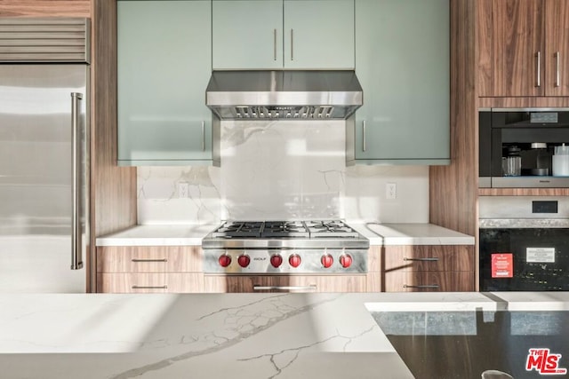 kitchen featuring stainless steel appliances, light stone counters, green cabinetry, decorative backsplash, and wall chimney exhaust hood