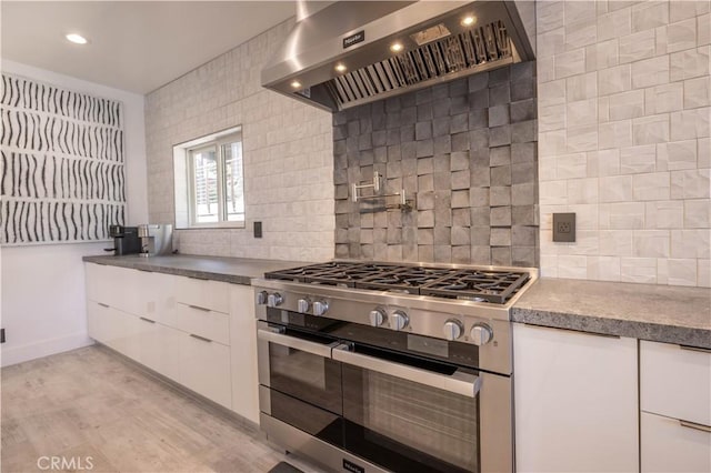 kitchen with range with two ovens, wall chimney range hood, light hardwood / wood-style floors, decorative backsplash, and white cabinets