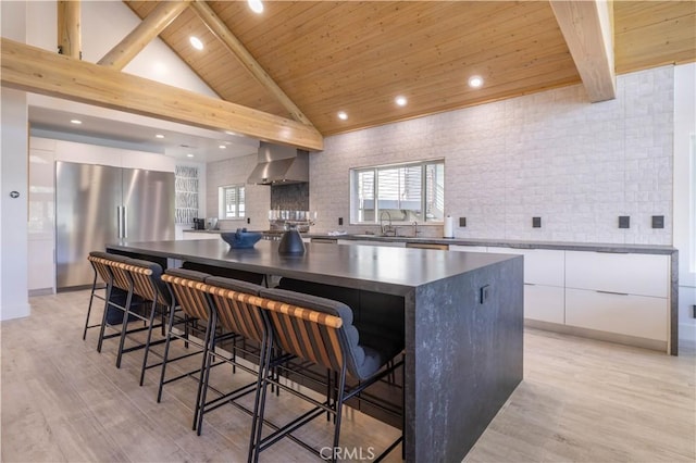 kitchen featuring wall chimney exhaust hood, white cabinetry, wood ceiling, stainless steel fridge, and a large island