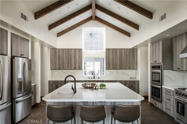 kitchen featuring beamed ceiling, appliances with stainless steel finishes, an island with sink, and decorative backsplash