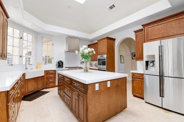 kitchen with sink, wall chimney range hood, stainless steel appliances, a tray ceiling, and a kitchen island