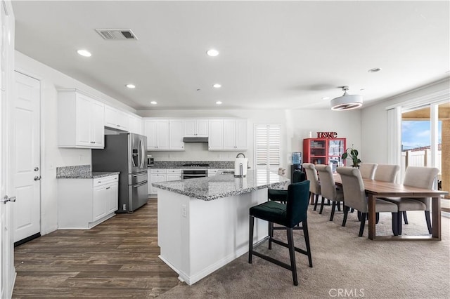 kitchen featuring light stone counters, stainless steel fridge with ice dispenser, an island with sink, white cabinets, and stove