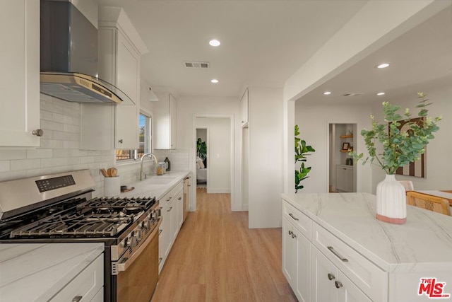 kitchen featuring wall chimney range hood, sink, light stone counters, white cabinets, and stainless steel range with gas cooktop