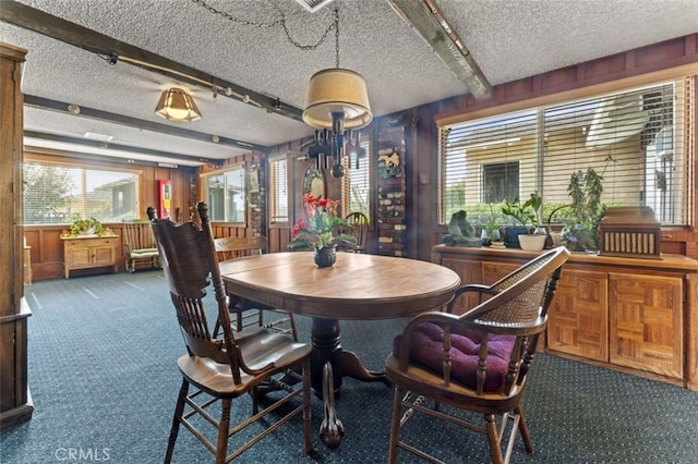 dining room with wooden walls, a textured ceiling, dark colored carpet, and beamed ceiling