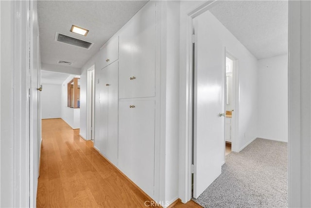 hallway with light wood-type flooring, visible vents, light carpet, and a textured ceiling