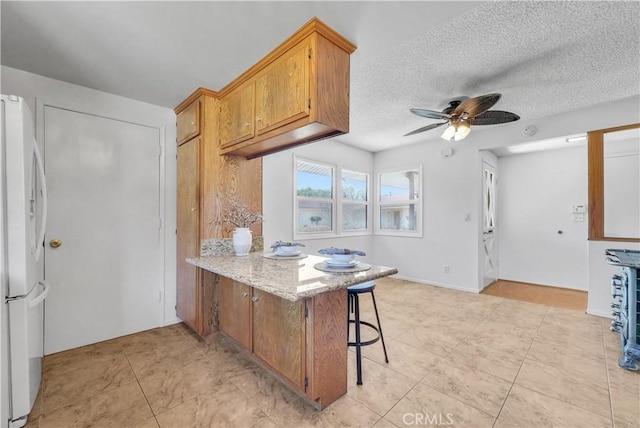 kitchen with ceiling fan, a peninsula, freestanding refrigerator, brown cabinetry, and a textured ceiling