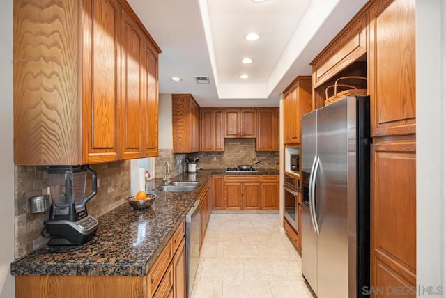 kitchen with sink, dark stone countertops, stainless steel appliances, tasteful backsplash, and a tray ceiling