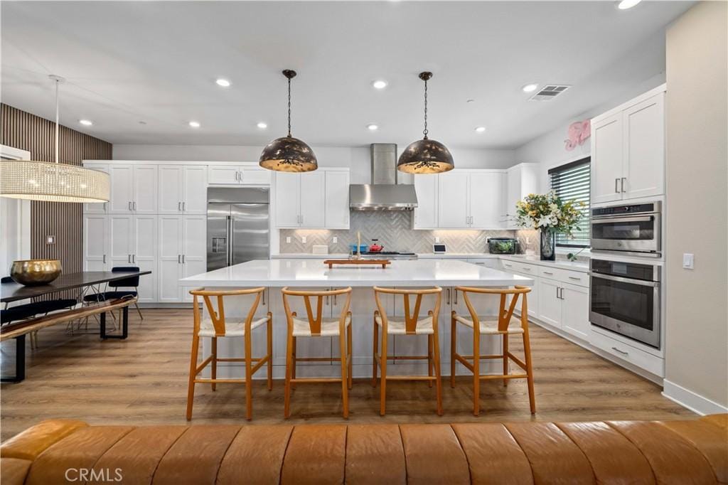 kitchen with appliances with stainless steel finishes, decorative light fixtures, white cabinets, wall chimney range hood, and a center island with sink