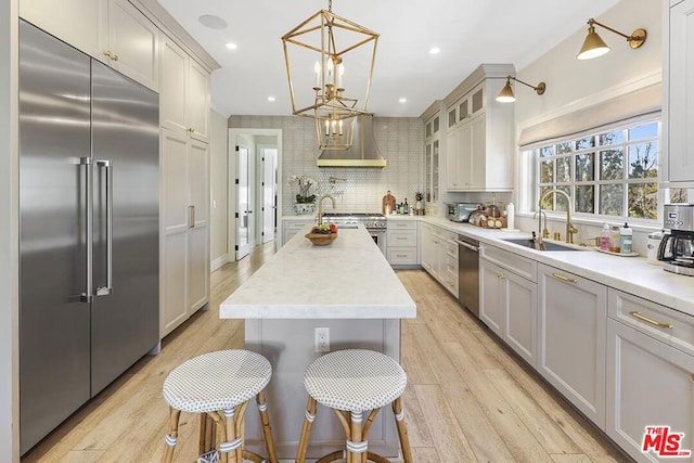 kitchen featuring high end appliances, decorative light fixtures, light wood-type flooring, an island with sink, and wall chimney range hood