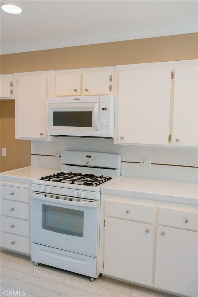 kitchen with tile countertops, white cabinets, decorative backsplash, crown molding, and white appliances