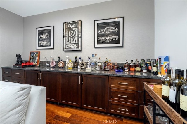 bar with dark wood-type flooring, sink, dark brown cabinetry, and dark stone counters