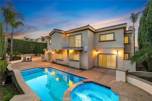 back house at dusk with exterior kitchen, an in ground hot tub, a balcony, and a patio