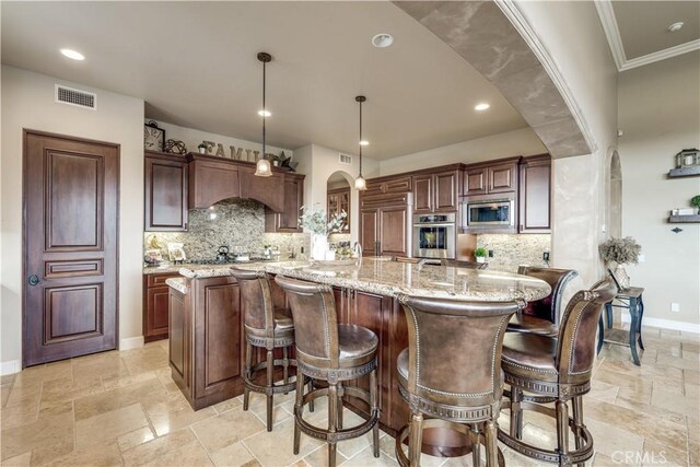 kitchen featuring light stone counters, built in appliances, decorative light fixtures, a large island with sink, and backsplash