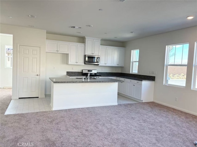 kitchen with white cabinetry, stainless steel appliances, a center island with sink, light carpet, and dark stone counters