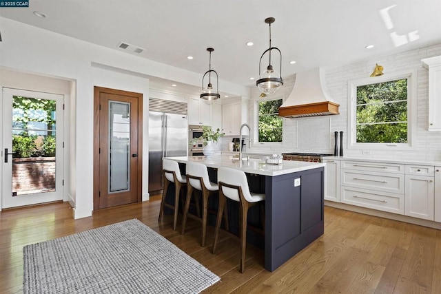 kitchen with premium range hood, a kitchen island with sink, white cabinetry, appliances with stainless steel finishes, and decorative light fixtures