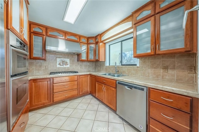 kitchen with brown cabinetry, a sink, under cabinet range hood, glass insert cabinets, and stainless steel appliances