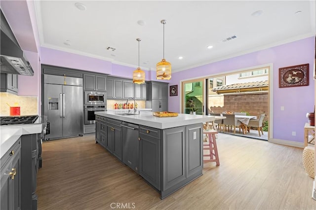 kitchen featuring wall chimney exhaust hood, sink, built in appliances, decorative light fixtures, and a kitchen island with sink