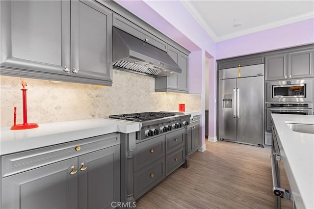 kitchen with appliances with stainless steel finishes, ornamental molding, decorative backsplash, wall chimney exhaust hood, and light wood-type flooring