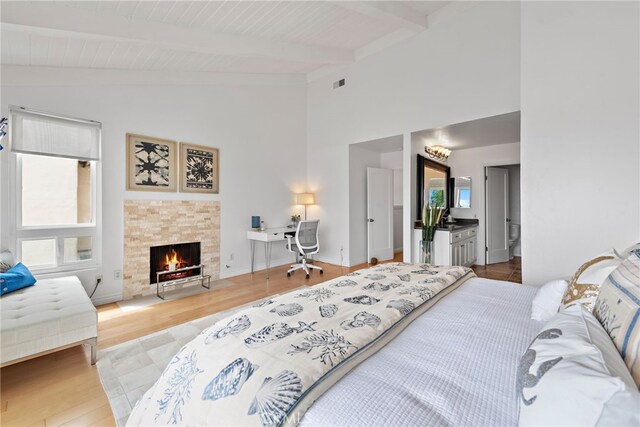 bedroom featuring connected bathroom, a stone fireplace, lofted ceiling with beams, and light wood-type flooring