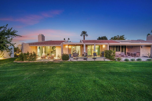 back house at dusk featuring a patio area and a lawn