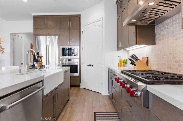 kitchen featuring sink, decorative backsplash, stainless steel appliances, wall chimney range hood, and light wood-type flooring