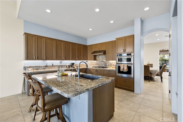 kitchen featuring stone countertops, a kitchen breakfast bar, stainless steel double oven, under cabinet range hood, and gas stovetop