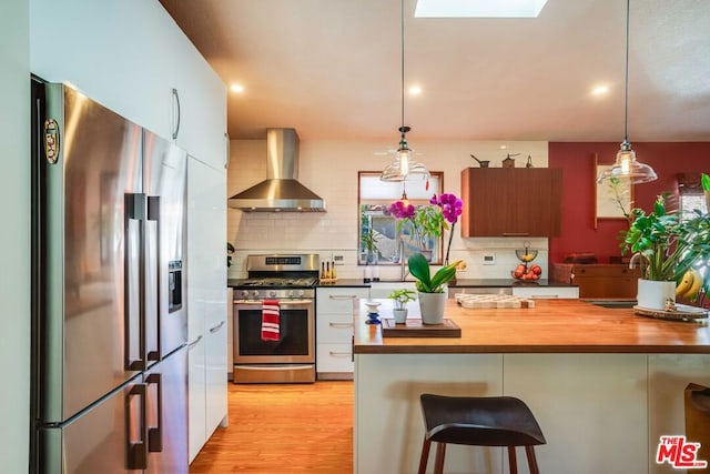 kitchen featuring wall chimney exhaust hood, wooden counters, decorative light fixtures, appliances with stainless steel finishes, and white cabinets