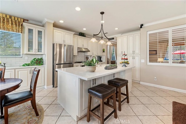 kitchen featuring light tile patterned flooring, decorative light fixtures, stainless steel fridge, an island with sink, and backsplash