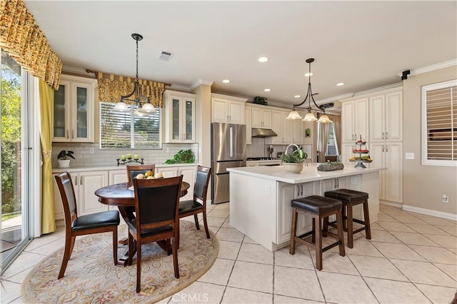 kitchen featuring light tile patterned flooring, stainless steel refrigerator, decorative light fixtures, an island with sink, and crown molding