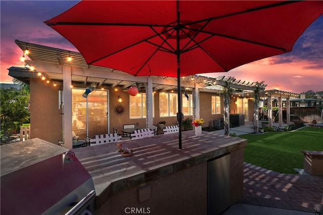 patio terrace at dusk with grilling area, a pergola, a lawn, and an outdoor kitchen