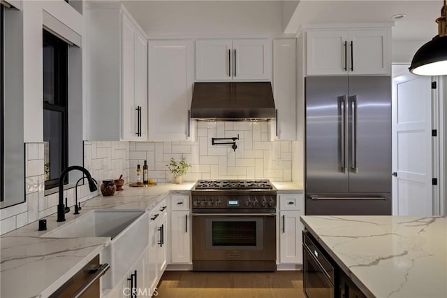 kitchen with white cabinetry, high quality appliances, light stone counters, tasteful backsplash, and exhaust hood