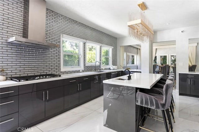 kitchen featuring wall chimney exhaust hood, a kitchen bar, stainless steel gas cooktop, a center island with sink, and decorative backsplash