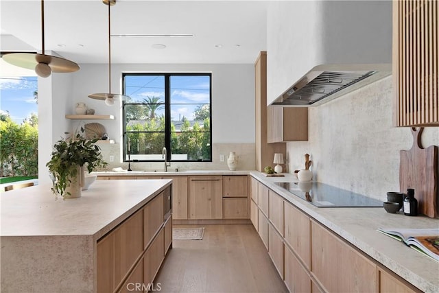 kitchen featuring wall chimney range hood, black electric stovetop, light brown cabinets, decorative light fixtures, and light wood-type flooring