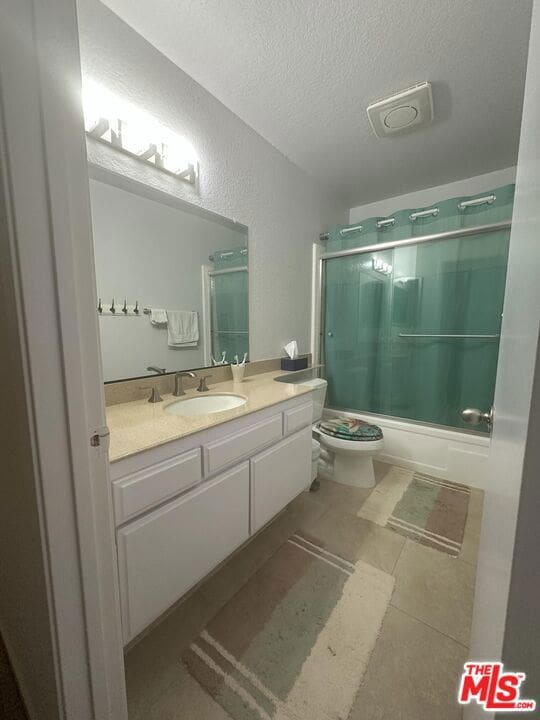 full bathroom with bath / shower combo with glass door, vanity, a textured ceiling, and toilet