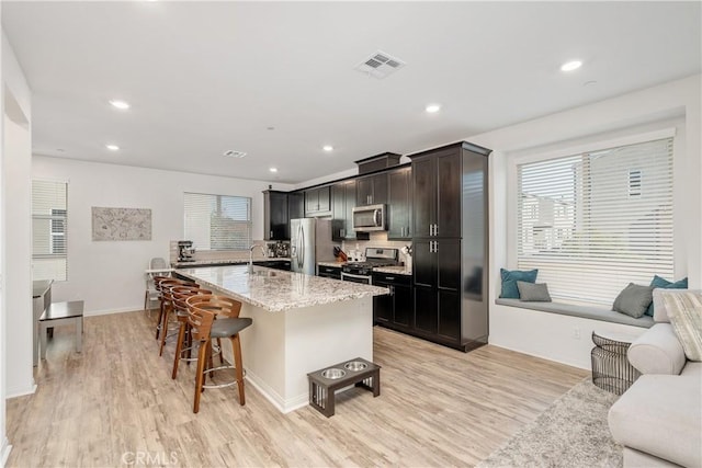 kitchen with light stone counters, a center island with sink, light wood-type flooring, appliances with stainless steel finishes, and a kitchen breakfast bar