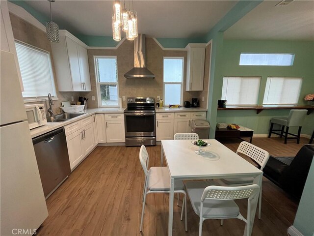 kitchen with wall chimney range hood, sink, hanging light fixtures, stainless steel appliances, and white cabinets