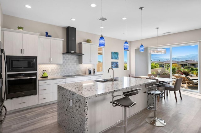 kitchen featuring white cabinetry, stainless steel oven, hanging light fixtures, a center island with sink, and wall chimney range hood