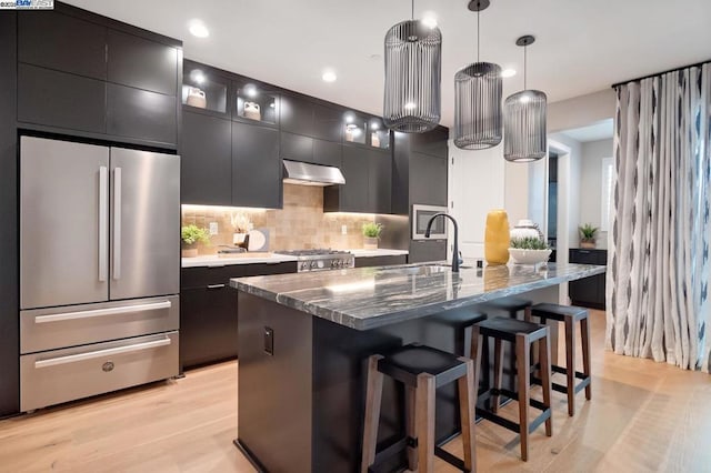 kitchen with sink, decorative light fixtures, light wood-type flooring, stainless steel refrigerator, and a kitchen island with sink