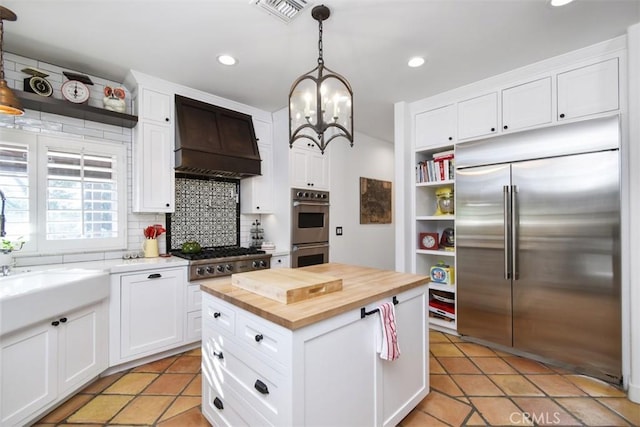 kitchen with premium range hood, appliances with stainless steel finishes, butcher block counters, white cabinets, and hanging light fixtures