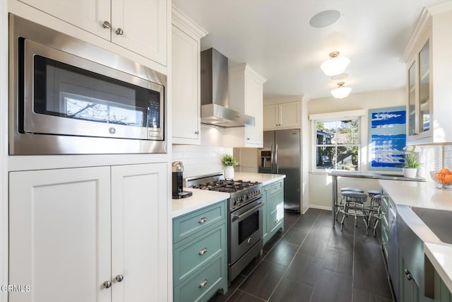 kitchen with wall chimney exhaust hood, tasteful backsplash, green cabinetry, stainless steel appliances, and white cabinets