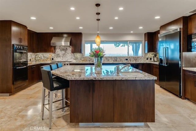 kitchen with light stone counters, wall chimney exhaust hood, an island with sink, and black appliances